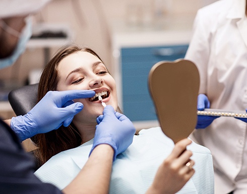 Dentist holding shade guide to patient’s teeth as she looks in mirror