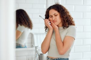 Woman in bathroom holding toothbrush and pressing hands to face in pain